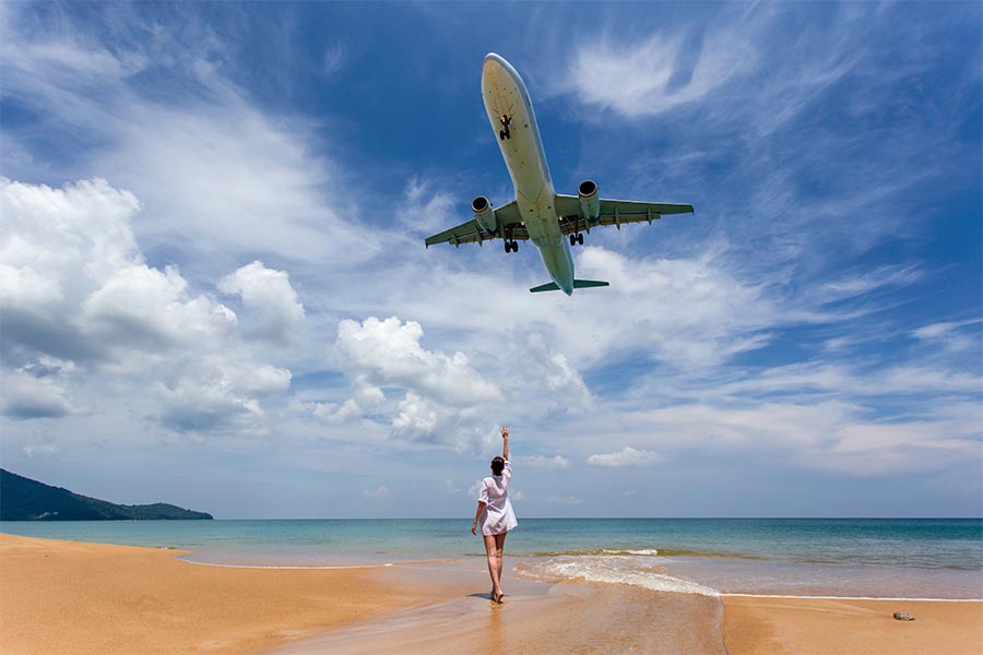 Una viajera en la playa de Mai Khao observa un avión aterrizando en Phuket, una imagen que ilustra los vuelos directos a Tailandia.